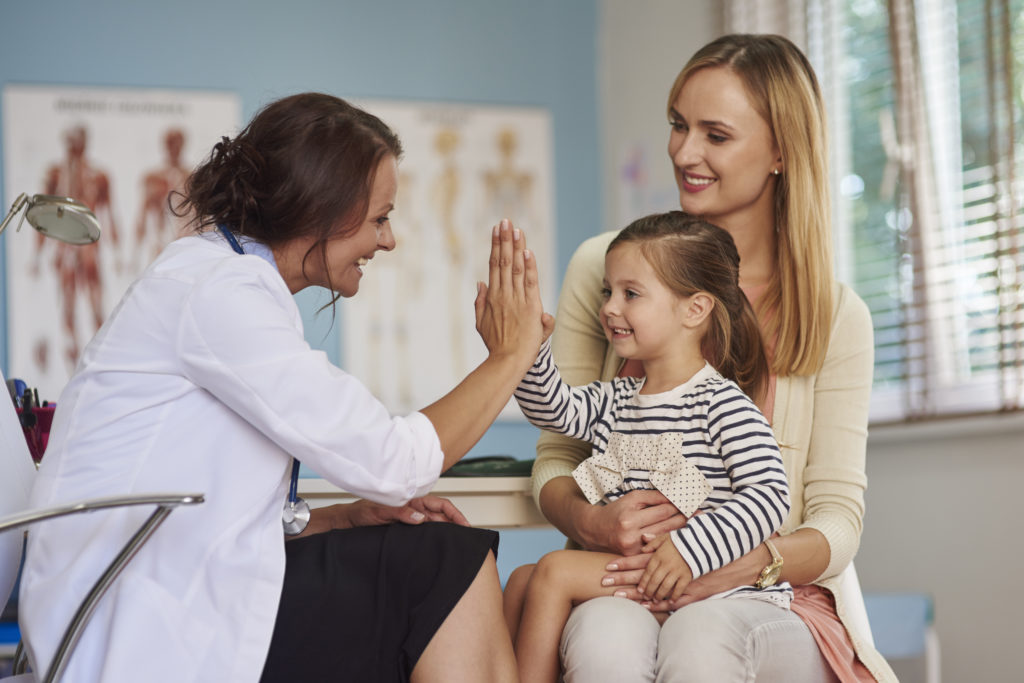 Female Physician high=ficing child patient who is sitting on her mother's lap in an exam room.